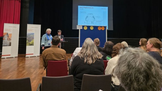 Das Foto zeigt im Vordergrund sitzendes Publikum von hinten und weiter hinten zwei Frauen, die zu einem Thema referieren.