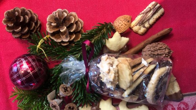 Das Bild zeigt eine Tüte Plätzchen, drumherum dekoriert mit Tannenzapfen, Kugeln und anderem Schmuck auf einer roten Tischdecke.